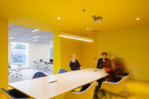 Nieuwbouw kantoor in Antwerpen door FRIDAY office for architecture. Vergaderruimte met felle gele muren en plafond. Achter de vergadertafel is een raam met uitzicht op de achterliggende kantoorruimte.