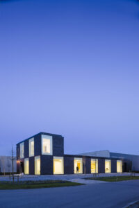 Nieuwbouw kantoor in Antwerpen door FRIDAY office for architecture. Nachtbeeld van een volume in zwarte baksteen met ritmische sequentie van raamopeningen. Het volume is vooraan dubbelhoog. Uit de ramen schijnt een warm licht. Door de ramen is het interieur zichtbaar.