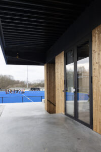Nieuwbouw hockeyclub voor Royal victory Hockey in Edegem door Friday Office for architecture. Glazen deur met zwarte kader in een houten buitengevel. Bovenaan is een overkraging te zien die dienst doet als luifel. Op de achtergrond het hockeyveld.