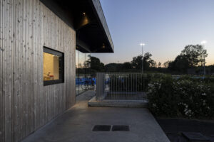 Nieuwbouw hockeyclub voor Royal victory Hockey in Edegem door Friday Office for architecture. Houten gevel met raam waarachter licht brandt. De gevel wordt overkraagd door het zwarte dak dat dienst doet als luifel.