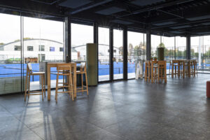 Nieuwbouw hockeyclub voor Royal victory Hockey in Edegem door Friday Office for architecture. Een interieurbeeld vanuit de cafetaria. Grote ramen geven uit op het hockeyveld. Voor de ramen staan hoge, houten tafels met houten krukken.