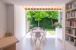 Uitbreiding van een gezinswoning. Een nieuwe uitbouw vloeit naadloos over in de bestaande woning. Hout tegen het plafond en in het meubilair zorgen voor de warme accenten. Door het driedelige schuifraam kan de tuin het huis maximaal binnen vloeien. Zicht vanuit de leefruimte door het schuifraam naar buiten toe.