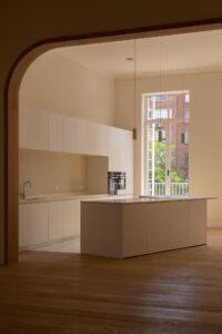 FRIDAY office deed een totaalrenovatie van die neoclassisistische pand in Antwerpen. Moderne, minimalistische keuken met witte kasten en een kookeiland, verlicht door natuurlijk licht via een groot raam met uitzicht op een bakstenen gebouw en groene bomen.