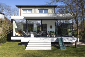Deze vrijstaande woning in interbellumstijl krijgt een beglaasde uitbouw. Geïnspireerd op de horizontale detaillering in de voorbouw, is de nieuwe annex opgevat als plateaus die het gebouw omarmen. Ze dragen de leefruimte, keuken en badkamer, afgewerkt in glas en zwart gebeitst hout. Het ruime uitkragende overdekt terras geeft beschutting voor een heerlijk English breakfast in het groen.