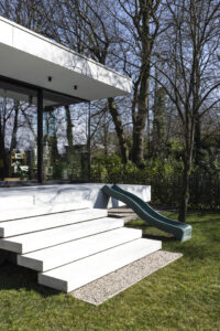 Deze vrijstaande woning in interbellumstijl krijgt een beglaasde uitbouw. Geïnspireerd op de horizontale detaillering in de voorbouw, is de nieuwe annex opgevat als plateaus die het gebouw omarmen. Ze dragen de leefruimte, keuken en badkamer, afgewerkt in glas en zwart gebeitst hout. Het ruime uitkragende overdekt terras geeft beschutting voor een heerlijk English breakfast in het groen.
