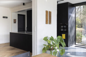 Deze vrijstaande woning in interbellumstijl krijgt een beglaasde uitbouw. Geïnspireerd op de horizontale detaillering in de voorbouw, is de nieuwe annex opgevat als plateaus die het gebouw omarmen. Ze dragen de leefruimte, keuken en badkamer, afgewerkt in glas en zwart gebeitst hout. Het ruime uitkragende overdekt terras geeft beschutting voor een heerlijk English breakfast in het groen. Dit is een interieurfoto.