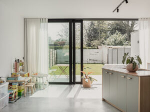 renovatie van een gezinswoning door friday office for architecture.De achtergevel op het gelijkvloers wordt compleet beglaasd, om de relatie met de tuin te vergroten. Op de verdieping blijft het volume gesloten. De nieuwe buitengevel werd geïsoleerd en afgewerkt met grijze leien. Deze interieurfoto toont het zicht vanuit de leefruimte, door het schuifraam, naar het terras toe.