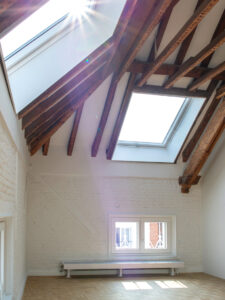 Verbouwing neoclassicistisch herenhuis door Friday Office for Architecture: lichte zolderruimte met witgeschilderde bakstenen muren, houten dakconstructie met zichtbare balken en twee grote dakramen waar zonlicht doorheen valt.”