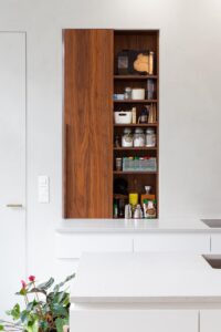 Renovatie van een gezinswoning door friday office for architecture. Detailfoto van het kruidenkastje in de keuken. Het kruidenkastje werd ingebouwd naast een raam dat uitgeeft op de inkomhal. Wanneer de deur van het kastje opengeschoven wordt, wordt het raam afgedekt door het deurtje.