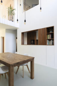 Intern worden de keuken en eetkamer gescheiden van de woonkamer. De woonkamer blijft op de eerste verdieping en krijgt aan de straatkant een groene buffer in de vorm van een uitkragende plantenbak.De keuken en eetkamer verhuizen naar het gelijkvloers en worden met een groot schuifraam verbonden met de tuin. De verdiepingsvloer boven de eetkamer verdwijnt en maakt plaats voor een royale vide. Deze ingreep zorgt voor lichtinval tot diep in de woning, waardoor ook een interne interactie tussen de woonkamer en eetkamer mogelijk blijft