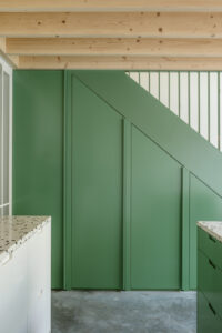 Moderne interieurarchitectuur met een open keuken en grote glazen pui die tuin en leefruimte naadloos verbindt. De combinatie van groene accenten, houten details en overvloedig daglicht creëert een warme, natuurlijke sfeer in deze eigentijdse woninguitbreiding.