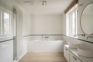 Renovatie van een gezinswoning door friday office for architecture. Een lichte, witte badkamer wordt gekenmerkt door een groene strip in gelakte tegel, die de omtrek van de ruimte volgt.